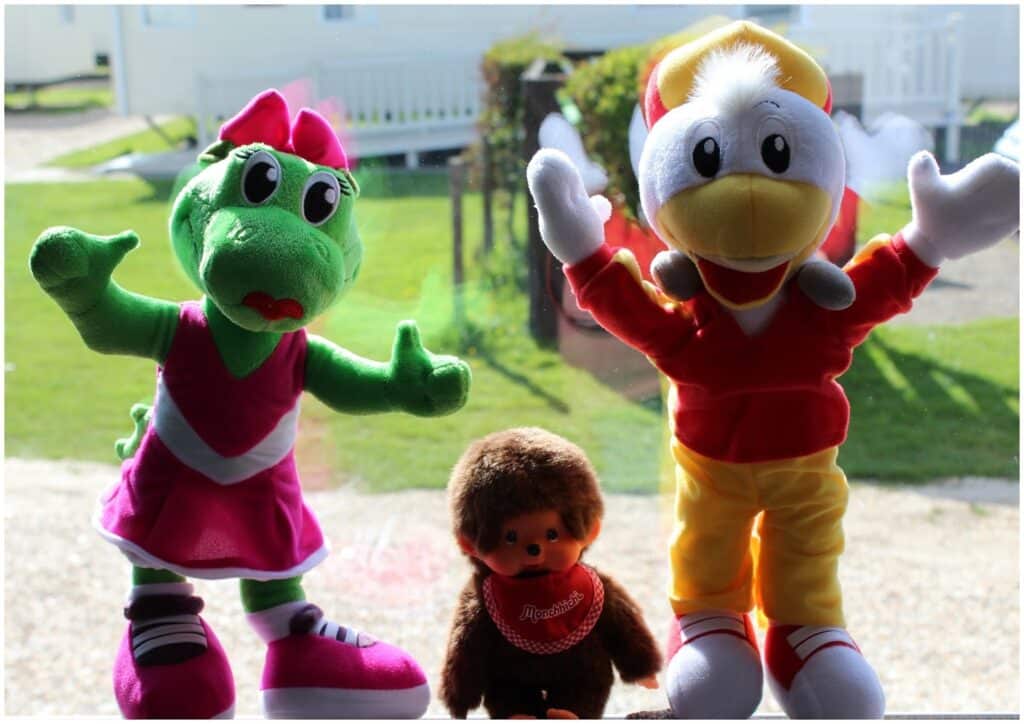 Three soft toys are displayed. On the left is a green dinosaur in a pink dress and bow. In the middle is a small brown monkey with a red bib. On the right is a white duck wearing a red and yellow outfit and a cap. All are standing with arms raised.