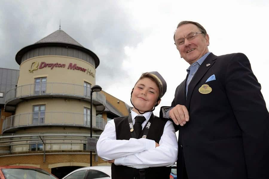A smiling boy in a hotel uniform and an older man in a suit stand outside Drayton Manor Hotel. The boy looks confident with arms folded, wearing a cap and waistcoat. The man, wearing glasses and a name badge, smiles beside him. The hotel building is in the background.