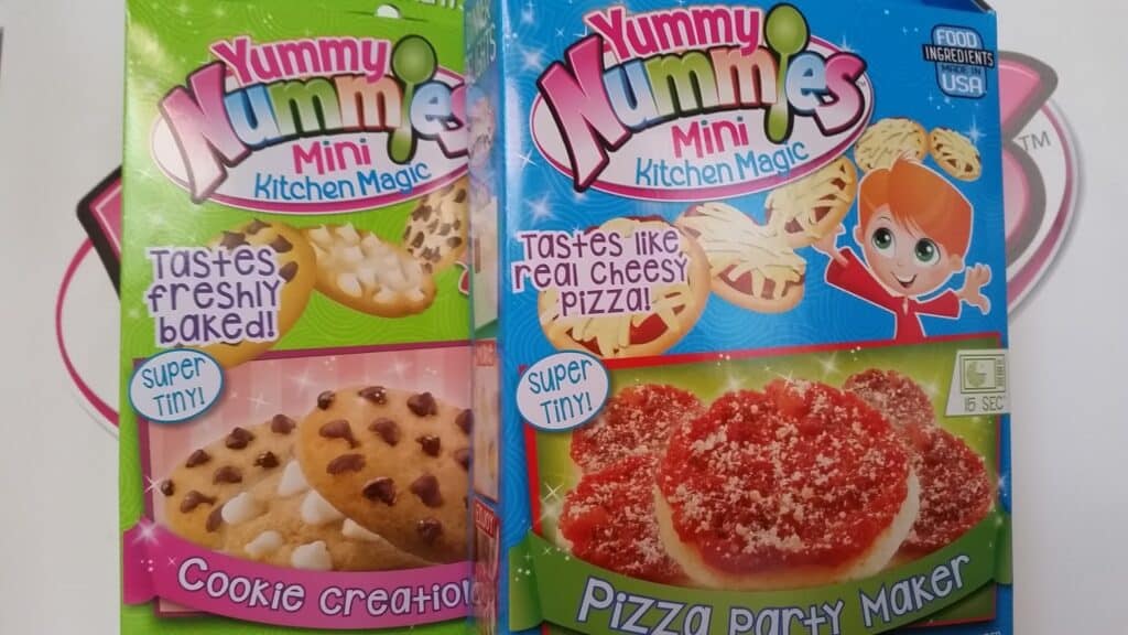 Two colourful Yummy Nummies Mini Kitchen Magic kits. The left box shows biscuit creations with chocolate chips, labelled “Tastes freshly baked!” The right box shows mini pizzas, labelled “Tastes like real cheesy pizza!” and features a cartoon chef girl.