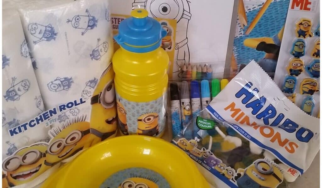 A collection of Minions-themed items, including kitchen roll, a yellow water bottle, bowl, Haribo Minions gummy sweets, colouring pens, and activity sheets, all featuring yellow Minions characters from Despicable Me. The items are brightly coloured and playful.