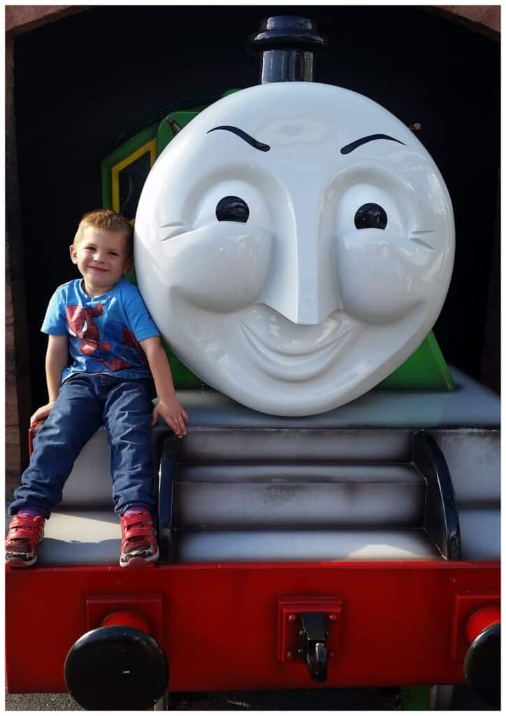 A young boy in a blue Spiderman T-shirt and red shoes sits smiling on the front of a large Thomas the Tank Engine character replica, with the character’s big white face and cheerful expression visible behind him.