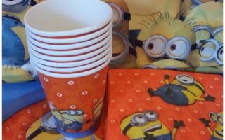 A party set-up with Minions-themed tableware: stacked cups, plates, and serviettes. All items feature yellow Minions characters on a red background with small white circles. The backdrop also displays Minions faces, creating a playful, animated scene.