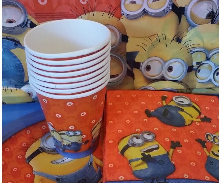A party set-up with Minions-themed tableware: stacked cups, plates, and serviettes. All items feature yellow Minions characters on a red background with small white circles. The backdrop also displays Minions faces, creating a playful, animated scene.