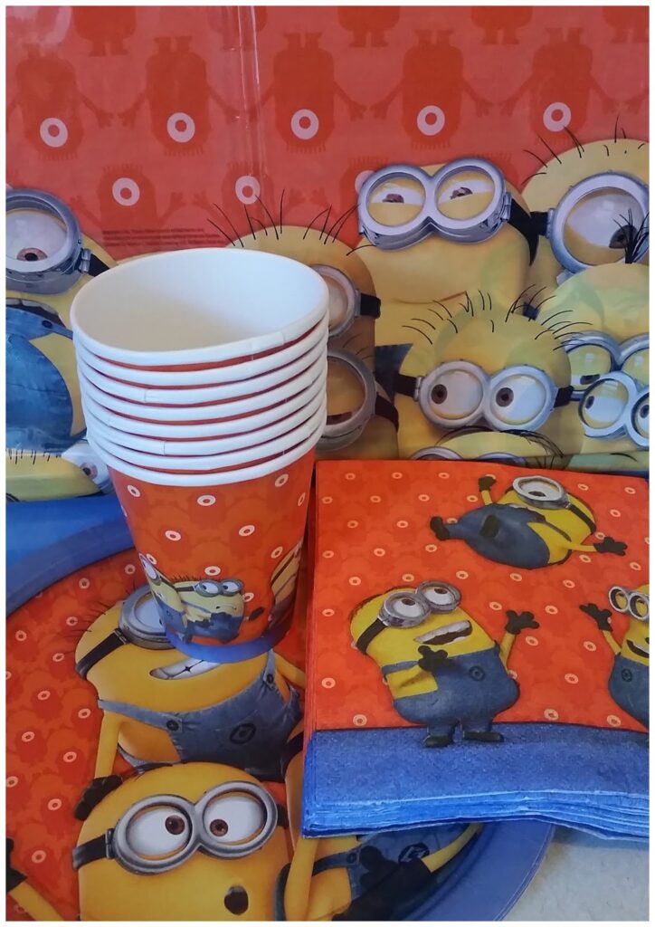 A stack of paper cups and folded serviettes with Minions from "Despicable Me" on an orange background. Behind them is a backdrop also featuring Minions. The scene appears festive, likely set up for a themed party.