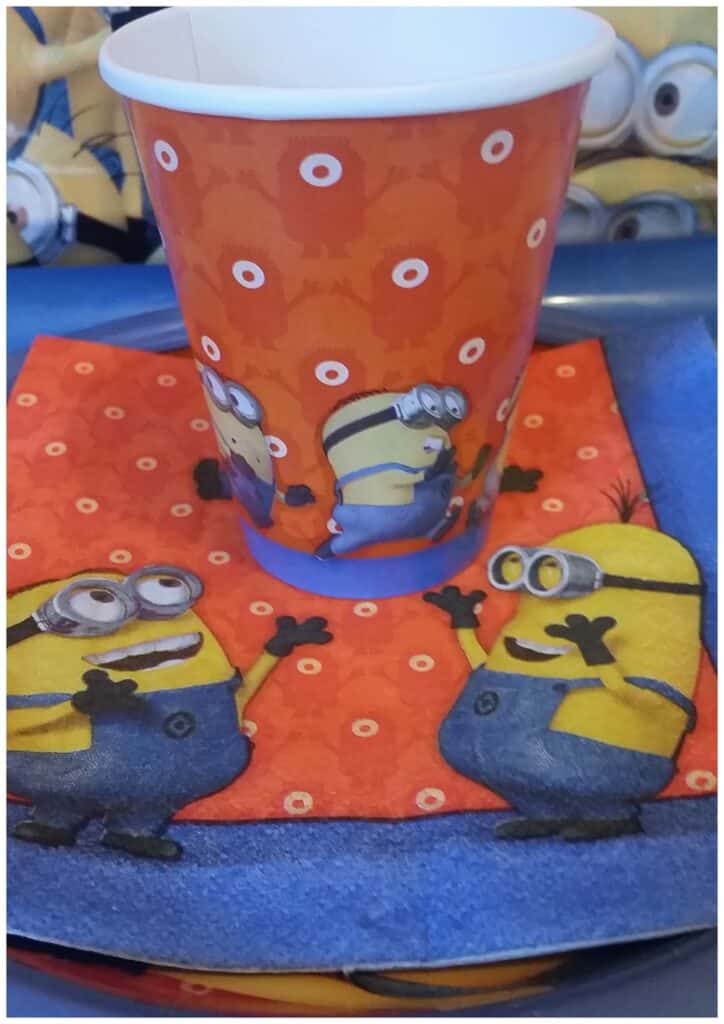 A paper cup and matching serviette feature Minions from the film "Despicable Me." The cup and serviette both have an orange background with small, white circles and illustrations of Minions in playful poses, set on a blue tablecloth.