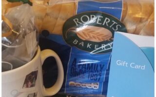 A loaf of Roberts Bakery white bread, a Tesco gift card, and a mug with a ribbon and small photos on it are grouped together on a white surface. The bakery packaging is blue and yellow, and the gift card is light blue with white writing.