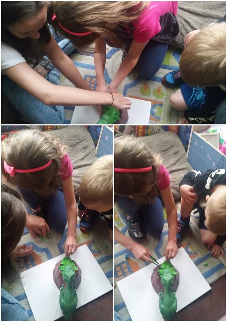 Three children sit on a colourful carpet, closely gathered around a green dinosaur model placed on white paper. Two girls carefully press green modelling clay onto the dinosaur's back, while a young boy watches with interest. The scene is captured from above.