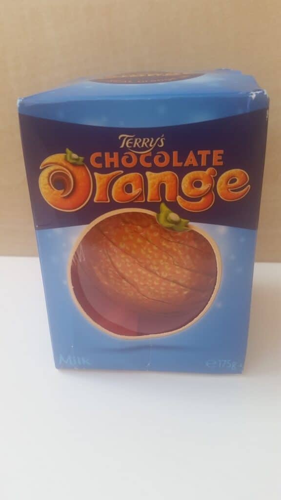 A blue 175g box of Terry’s Chocolate Orange, showing the brand name in orange and brown letters above an image of a chocolate orange, partially visible through a round window in the packaging. The word “Milk” appears at the bottom left.