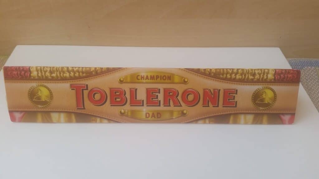 A Toblerone chocolate bar in a special box reading “CHAMPION TOBLERONE DAD” in gold and red text, decorated with gold coins and a curtain-like background, placed on a white surface against a wooden backdrop.