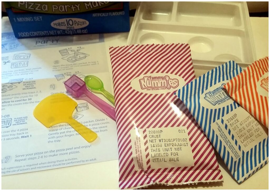 A close-up of a Yummy Nummies Pizza Party Maker kit shows striped ingredient packets, a yellow plastic spatula, a purple mixing spoon, a white mixing tray, and an instruction sheet with illustrated steps partially visible under the utensils.