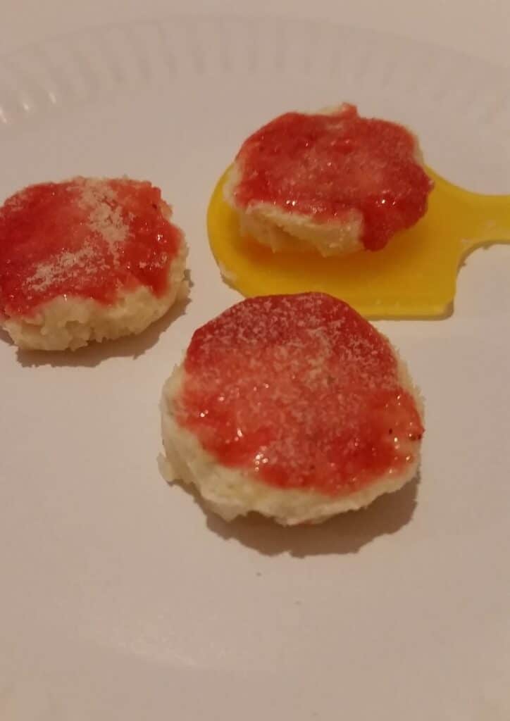 Three round biscuits topped with red jelly and a light dusting of white powder are on a white plate. One biscuit rests on a small yellow plastic paddle, while the others sit directly on the plate. The background is plain and neutral.