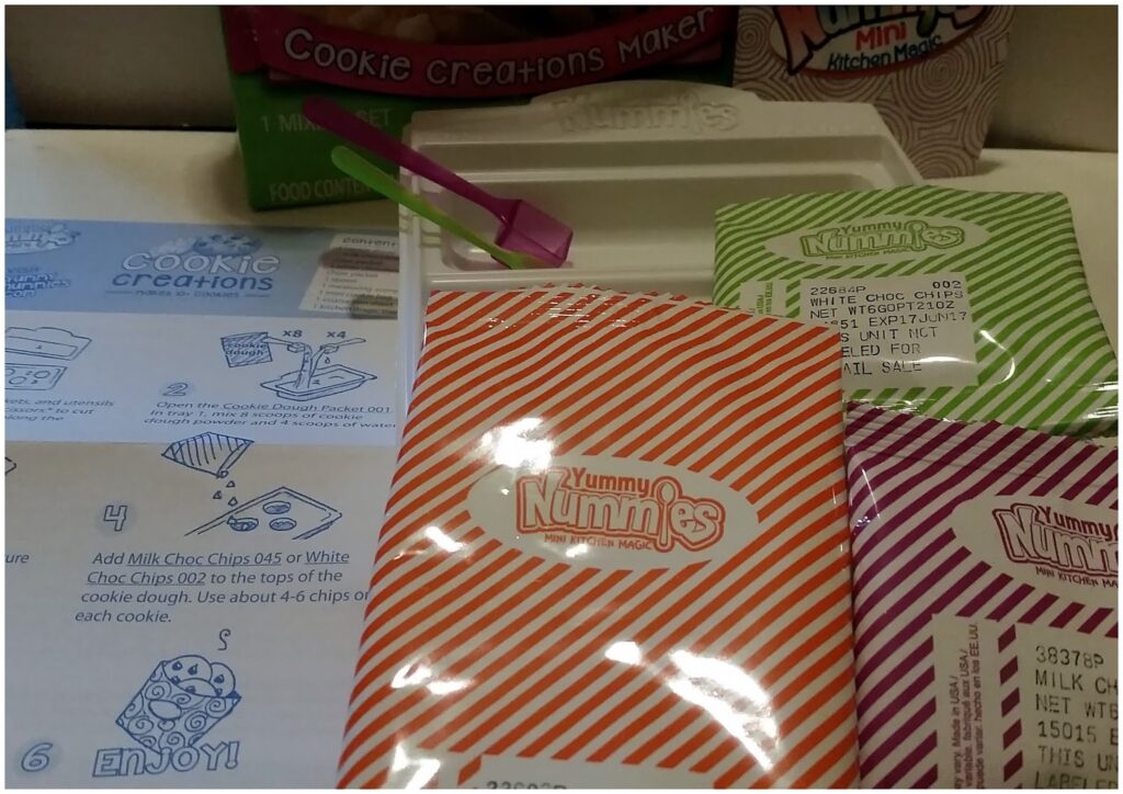 Close-up of a Yummy Nummies Cookie Creations Maker kit. Visible are colourful ingredient packets (orange, green, and purple stripes) labelled “Yummy Nummies,” part of the instruction sheet with illustrations, and a white plastic tray in the background.