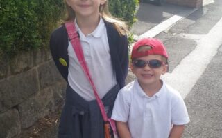 A young girl and a small boy stand on a sunny pavement. The girl wears a school uniform, jumper tied around her waist, and a pink shoulder bag. The boy wears a white shirt, black trousers, red cap back to front, and sunglasses. Both are smiling. Green bushes and houses are behind them.