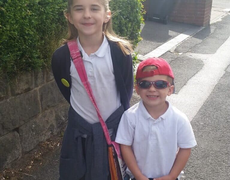 A young girl and a small boy stand on a sunny pavement. The girl wears a school uniform, jumper tied around her waist, and a pink shoulder bag. The boy wears a white shirt, black trousers, red cap back to front, and sunglasses. Both are smiling. Green bushes and houses are behind them.