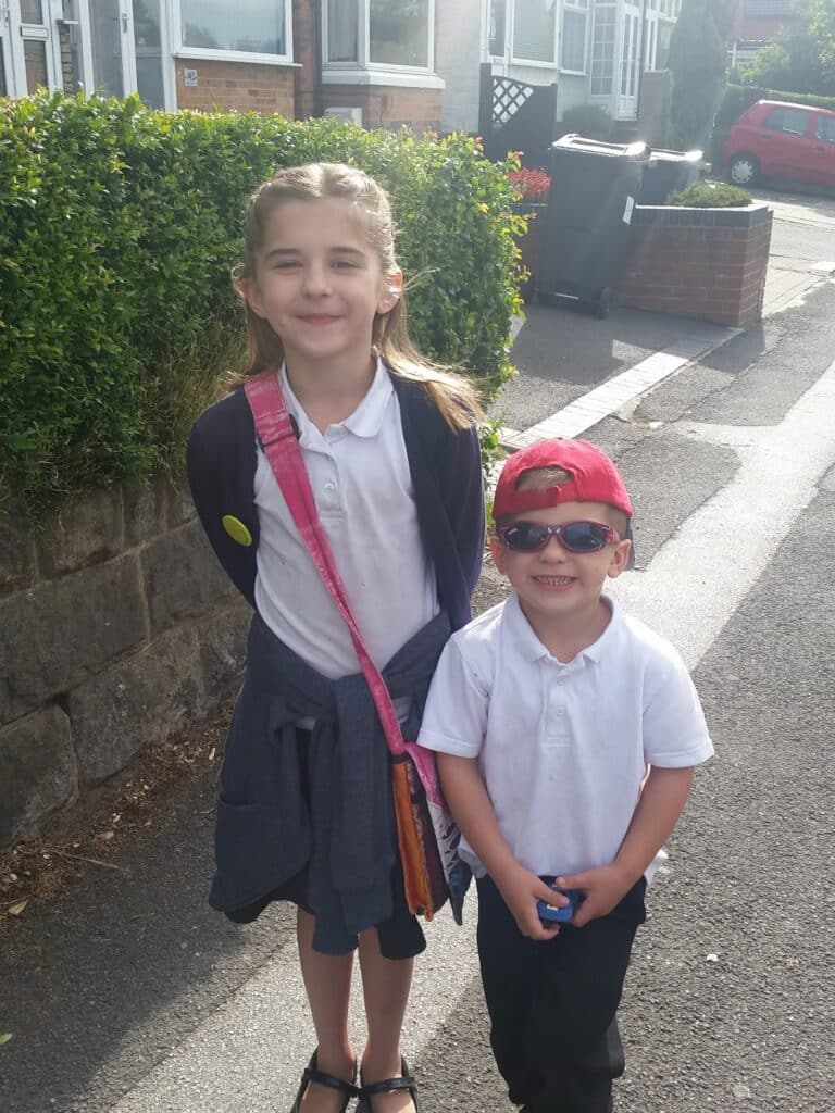 Two children stand outside on a sunny day. The older girl has long hair, wears a white shirt, dark skirt, and cardigan, with a pink shoulder bag. The younger boy wears a white shirt, dark trousers, red cap, and sunglasses, and is smiling widely.