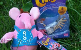 A pink Clangers plush toy with a blue waistcoat sits on green grass next to a packaged Clangers “Iron Chicken” figurine. Both items are Clangers merchandise, with the packaging displaying the logo and colourful design.