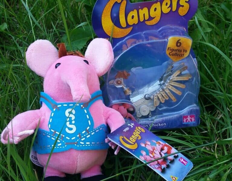 A pink Clangers plush toy with a blue waistcoat sits on green grass next to a packaged Clangers “Iron Chicken” figurine. Both items are Clangers merchandise, with the packaging displaying the logo and colourful design.