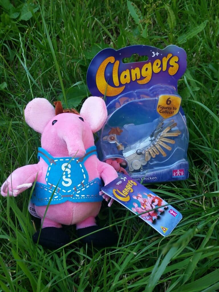 A pink Clangers soft toy wearing a blue outfit sits on green grass. Next to it is a packaged Clangers toy labelled “Iron Chicken,” featuring a bird figure with metallic feathers. Both items are Clangers merchandise.