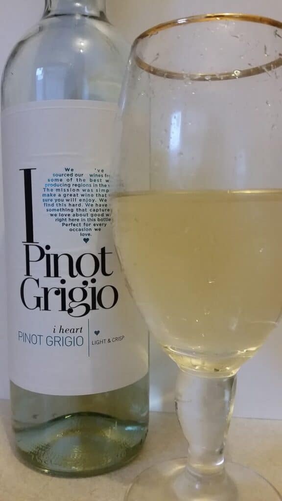 A clear wine glass filled with pale yellow pinot grigio stands in front of a bottle labelled "I heart Pinot Grigio." The bottle has a white label with black and blue text, and both items are placed on a light-coloured surface.