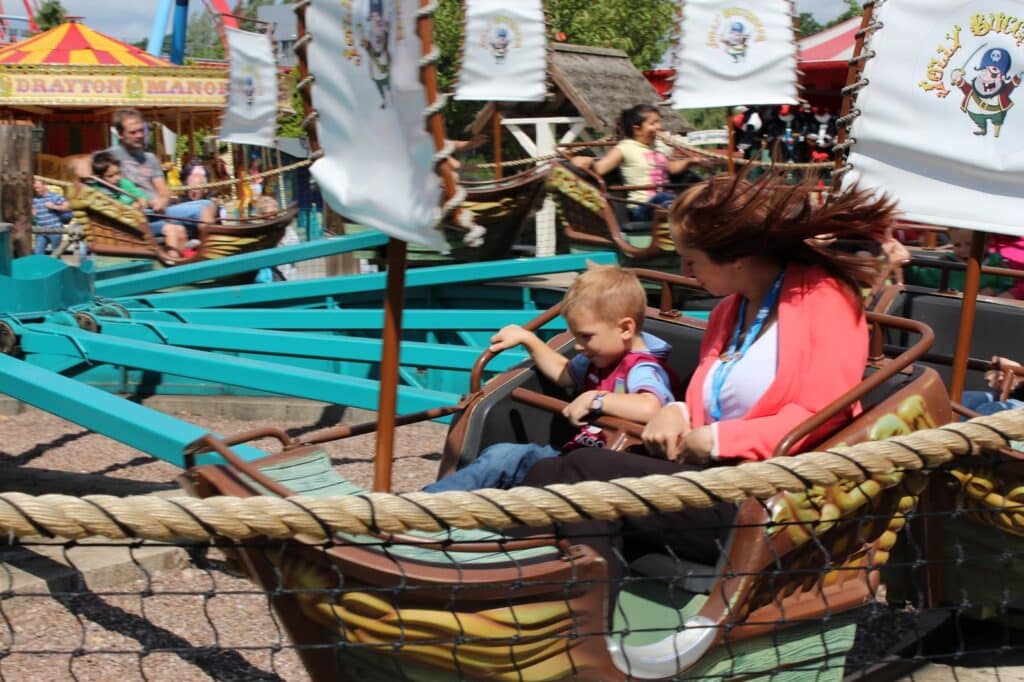 A woman and young boy ride a spinning amusement park ride. Both appear excited, with the woman’s hair blown back by the motion. Colourful banners and a “Drayton Manor” sign are visible in the background, along with other park visitors.