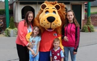 A woman and three children stand smiling with a person in a lion mascot costume outdoors. The woman wears a pink jacket, one girl wears a Frozen top, another wears a pink Adidas jacket, and the boy wears a football shirt. Buildings and greenery are in the background.