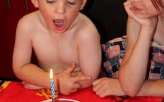 A bare-chested young boy excitedly looks at a lit birthday candle on a Spider-Man cake, with a girl beside him smiling and resting her chin on her hand. They’re seated at a table covered with a red cartoon-themed tablecloth.