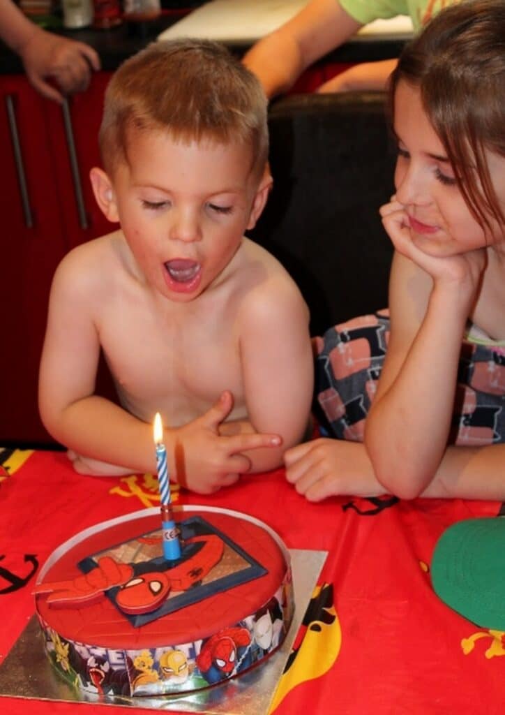 A young shirtless boy with short blond hair excitedly looks at a birthday cake with a single lit candle and Spider-Man decorations. A girl beside him, wearing patterned pyjamas, smiles and rests her chin on her hand, watching him. The tablecloth is red with cartoon designs.
