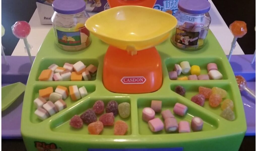 A colourful toy sweet shop set with a yellow-orange scale at the centre, jars, two red lollies, and trays holding various small, colourful toy sweets and gummies, arranged neatly in sections on a green and purple plastic tray.