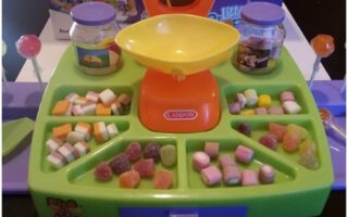 A colourful toy sweet shop set with a yellow-orange scale at the centre, jars, two red lollies, and trays holding various small, colourful toy sweets and gummies, arranged neatly in sections on a green and purple plastic tray.