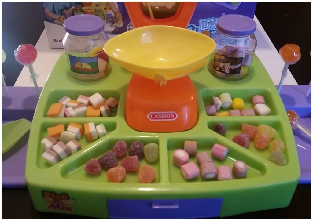 A colourful plastic toy sweet shop set with various compartments holding assorted toy sweets, a yellow and orange weighing scale in the centre labelled "CASDON," two jars, and four toy lollies, all arranged on a green and purple tray.