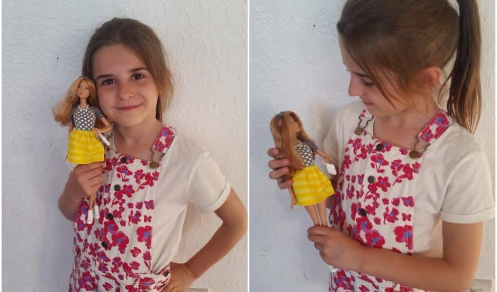 A young girl with brown hair, wearing a white shirt and pink floral dungarees, stands against a white textured wall. She smiles whilst holding a Barbie doll dressed in a grey polka-dot top and bright yellow skirt, looking at the doll in the second photo.