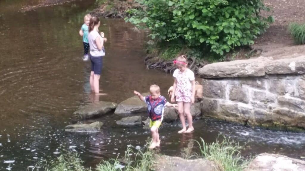 Three children and an adult stand in a shallow stream near large rocks and a stone wall. Two children are playing in the water, while one child and the adult stand near the edge. Lush green bushes grow nearby, and the scene looks relaxed and summery.