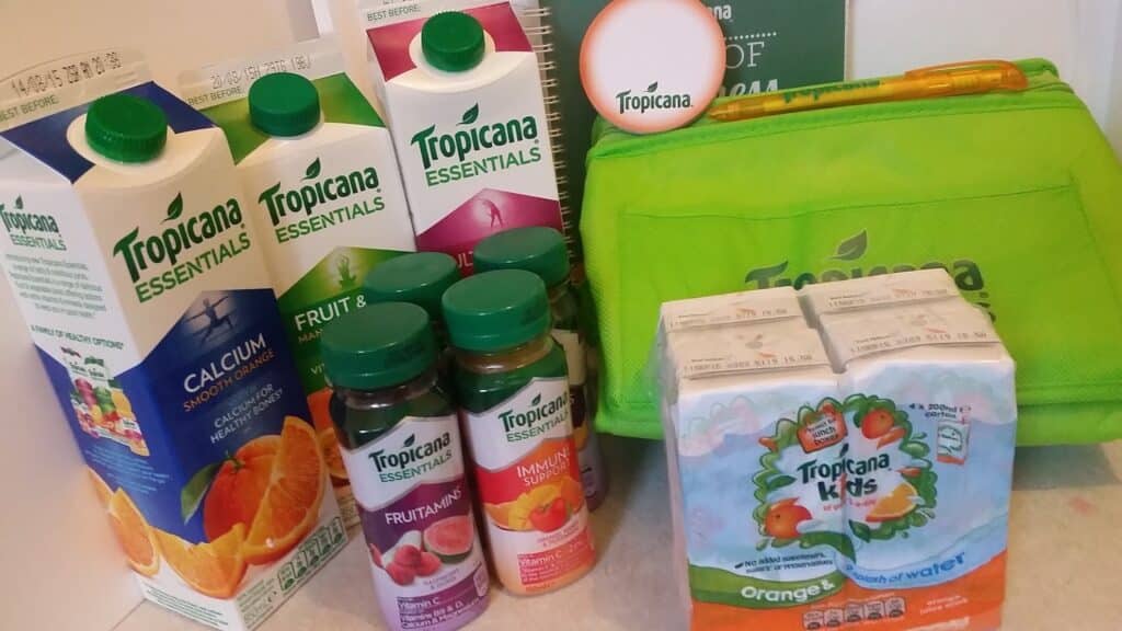 A selection of Tropicana juice products, including cartons, bottles, and juice cartons, are arranged next to a bright green insulated Tropicana lunch bag, a pen, and a round badge. The products display various flavours and colourful fruit imagery.