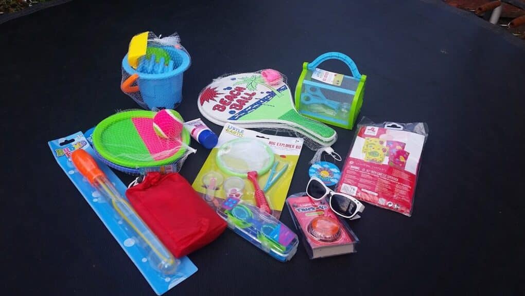 A collection of colourful kids’ summer toys and accessories on a dark surface, including racquets, a bubble wand, a sand bucket with tools, sunglasses, a skipping rope, a magnifying glass, and a small red pouch. Some items are still in their packaging.