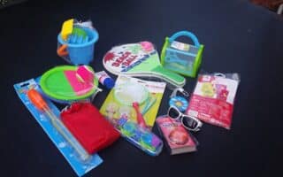 A collection of colourful kids’ summer toys and accessories on a dark surface, including racquets, a bubble wand, a sand bucket with tools, sunglasses, a skipping rope, a magnifying glass, and a small red pouch. Some items are still in their packaging.