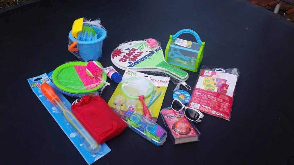 A collection of colourful beach and outdoor toys on a black surface, including a blue bucket with tools, toy racquets, a green net, bubbles, sunglasses, a red pouch, paddle ball sets, and various packaged summer items.