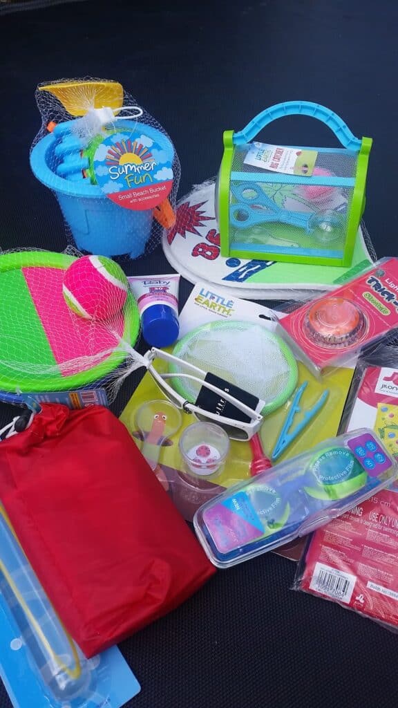 A variety of colourful summer toys and outdoor items, including a blue bucket with beach toys, a paddle ball set, skipping rope, bug-catching kit, frisbee, bubbles, red drawstring bag, sticky hand toy, Little Debbie cake, and more, arranged on a dark background.