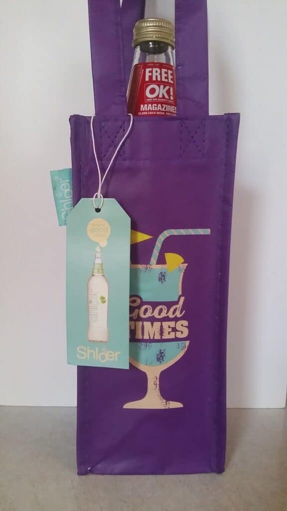 A purple gift bag with a cocktail glass illustration and the words "Good Times." A bottle with a red cap is inside, partly visible, with a tag that says "FREE! OK! MAGAZINE." A light blue tag with a white bottle and the word "Shloer" hangs from the bag.