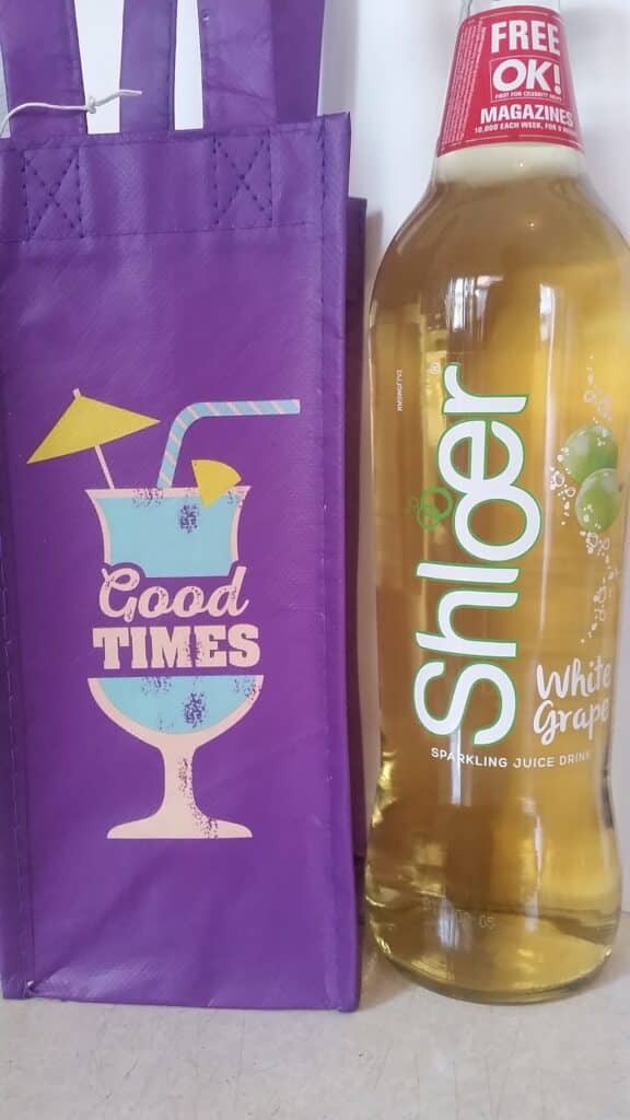 A purple bag with a cocktail glass, a straw, an umbrella, and the text "Good Times" stands next to a bottle of Shloer White Grape sparkling juice drink with a clear label and a red cap advertising "Free OK! Magazines.