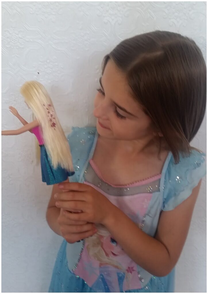 A young girl with straight brown hair, wearing a light blue dress with sparkles, looks down and smiles at a Barbie doll with long blonde hair and a blue skirt, which she is holding in front of a white textured wall.