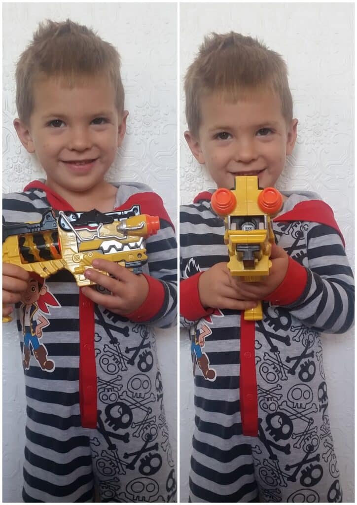 A young boy in striped pyjamas with skull and crossbones patterns smiles whilst holding a yellow and black toy, first showing its side and then pointing its front towards the camera, against a textured white wall.