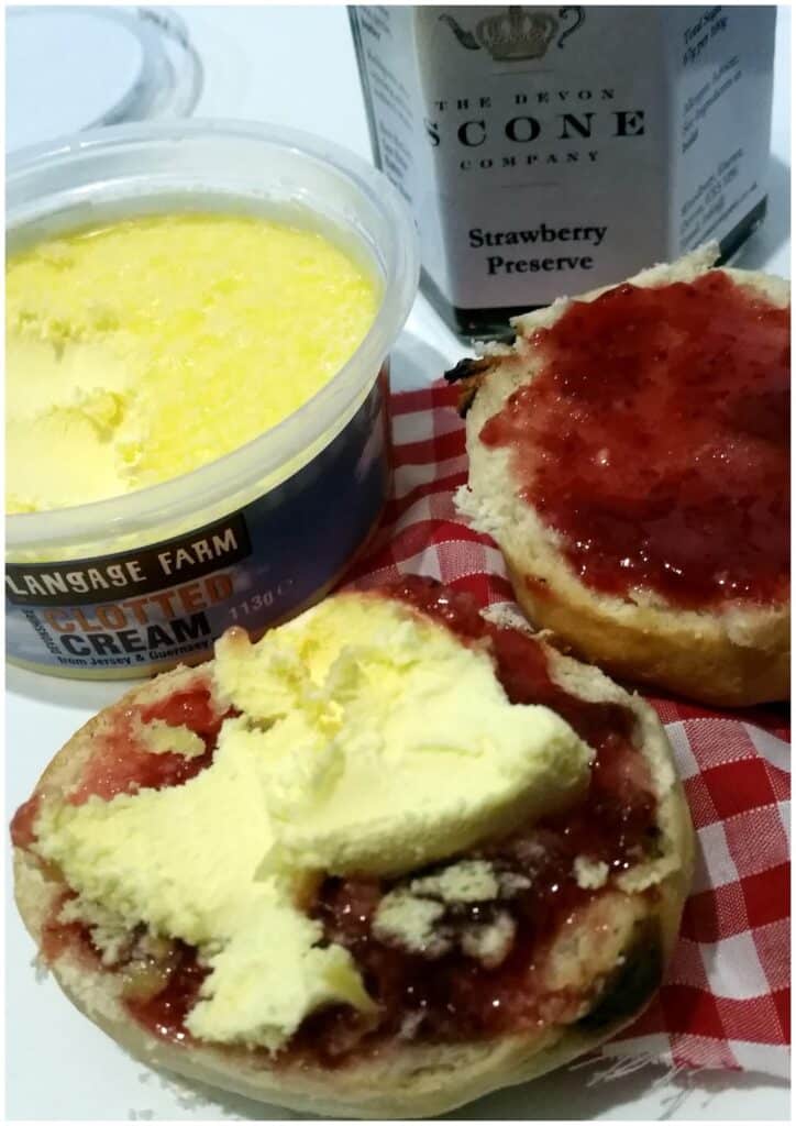 Two halves of a scone on a red chequered cloth—one spread with cream and strawberry jam, the other with just jam. Next to them are an open tub of clotted cream and a jar of strawberry preserve. The scene suggests a traditional cream tea.