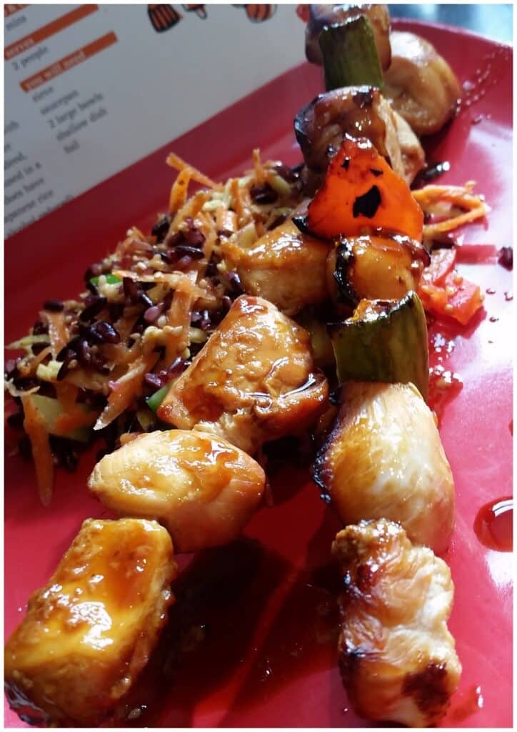 A close-up of glazed chicken and vegetable kebabs, featuring grilled chicken pieces, green courgette, and red pepper, on a red plate. In the background, there's a shredded vegetable salad and a partially visible recipe card.