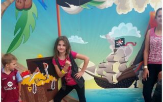 Three children stand in front of a colourful mural at Hatton Adventure World, featuring a pirate ship, treasure chest, and parrot. One girl strikes a confident pose, while a young boy and another girl stand nearby, all wearing casual clothes.