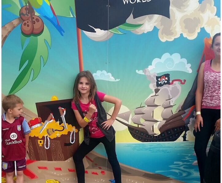Three children stand in front of a colourful mural at Hatton Adventure World, featuring a pirate ship, treasure chest, and parrot. One girl strikes a confident pose, while a young boy and another girl stand nearby, all wearing casual clothes.