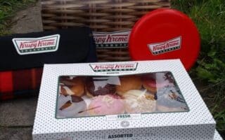 Two closed boxes of Krispy Kreme assorted doughnuts sit on the ground outdoors next to a branded tartan blanket, a red circular tin, and an open empty box. The setting is grassy with garden elements partially visible in the background.