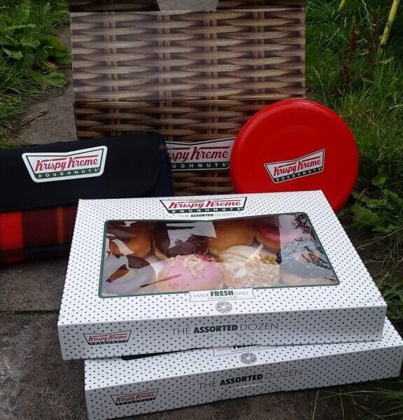 Two closed boxes of Krispy Kreme assorted doughnuts sit on the ground outdoors next to a branded tartan blanket, a red circular tin, and an open empty box. The setting is grassy with garden elements partially visible in the background.