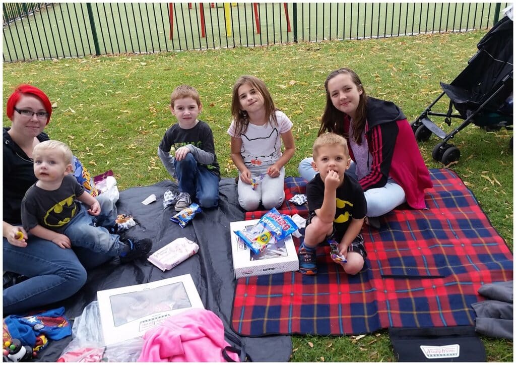 Six children and one adult sit on picnic blankets on grass, eating snacks. The adult, with red hair and glasses, sits to the left with a young blond boy. Nearby are boxes of food and snacks. A playground and black fence are in the background. It’s a cloudy day.