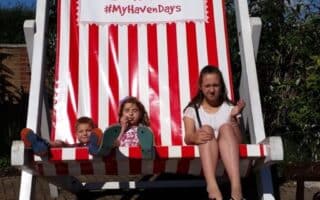 Three children sit on a giant red-and-white striped deckchair under a blue sky. The deckchair has a sign reading “Seashore #MyHavenDays.” The children smile and pose, with greenery and a brick path in the background.