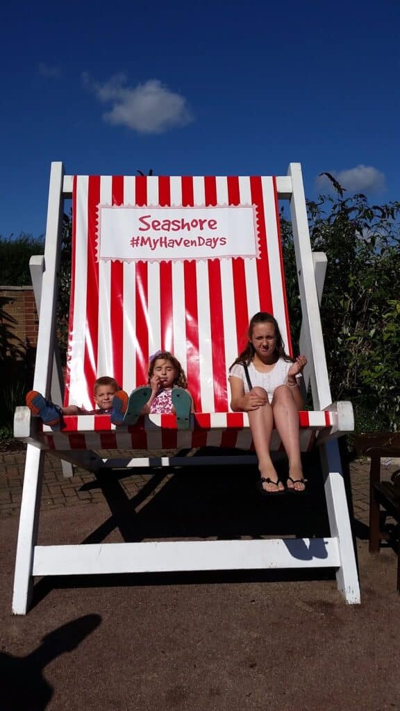 Three children sit on a giant red-and-white striped deckchair under a blue sky. The chair has a sign reading "Seashore #MyHavenDays." Two younger children lounge to the left, an older girl sits to the right with her legs dangling. Shrubs and paving are visible behind them.